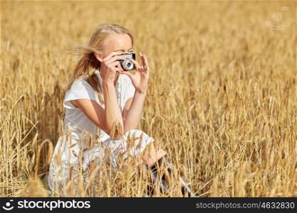 nature, summer holidays, vacation and people concept - happy young woman taking picture with vintage film camera in cereal field