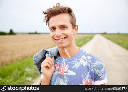 nature, summer, youth culture and people concept - smiling young hippie man on country road chewing rye spike
