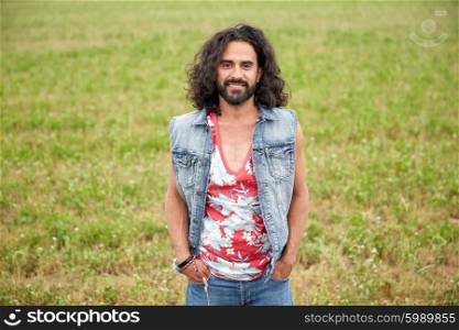 nature, summer, youth culture and people concept - smiling young hippie man in denim vest on green field