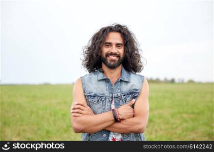 nature, summer, youth culture and people concept - smiling young hippie man in denim vest on green field