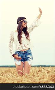 nature, summer, youth culture, gesture and people concept - smiling young hippie woman in sunglasses on cereal field waving hand