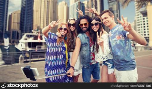 nature, summer, youth culture, technology and people concept - smiling hippie friends in sunglasses taking picture by smartphone on selfie stick and showing peace gesture over dubai city harbour