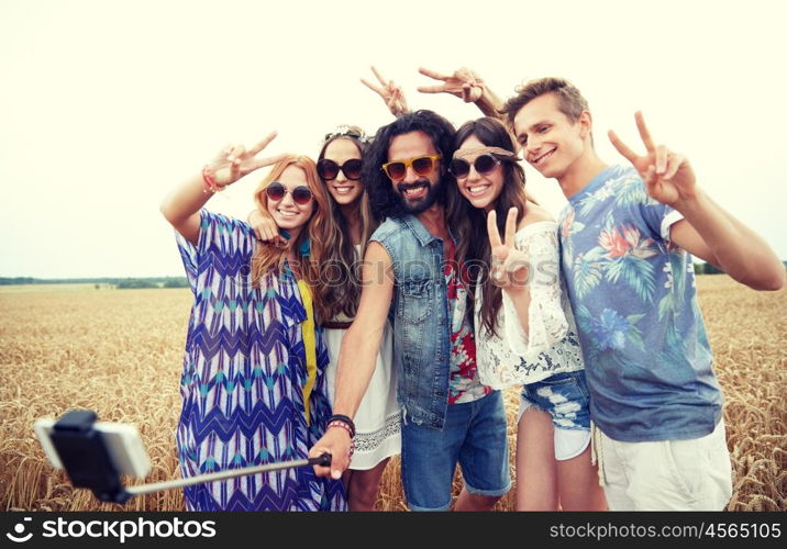 nature, summer, youth culture, technology and people concept - smiling young hippie friends in sunglasses taking picture by smartphone on selfie stick and showing peace gesture on cereal field