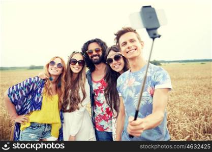 nature, summer, youth culture, technology and people concept - smiling young hippie friends in sunglasses taking picture by smartphone on selfie stick on cereal field