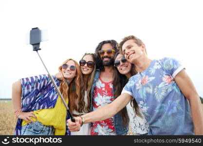 nature, summer, youth culture, technology and people concept - smiling young hippie friends in sunglasses taking picture by smartphone on selfie stick on cereal field