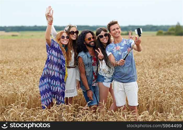 nature, summer, youth culture, technology and people concept - smiling young hippie friends in sunglasses taking picture by smartphone on selfie stick and showing peace gesture on cereal field