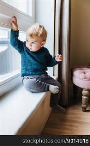 Naughty small kid with blonde hair and blue eyes, sits on window sill, looks out of window, notices something in yard. Curious cute child plays alone, looks outside with attentive expression