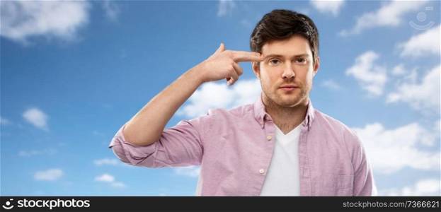 negative emotion, expression and stress concept - bored young man shooting himself to head by finger gun gesture over blue sky and clouds background. bored man shooting himself by finger gun gesture
