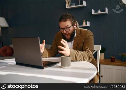 Negative frustrated guy in jacket and shorts shouting on laptop screen during company online meeting. Negative frustrated guy shouting on laptop screen
