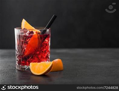 Negroni cocktail in crystal glass with orange slice and black straw on black table background.