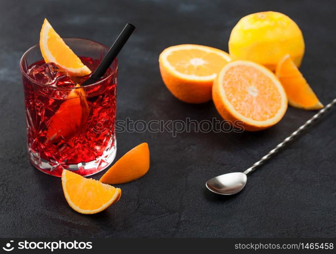 Negroni cocktail in crystal glass with orange slice and fresh raw oranges with spoon on black background. Top view