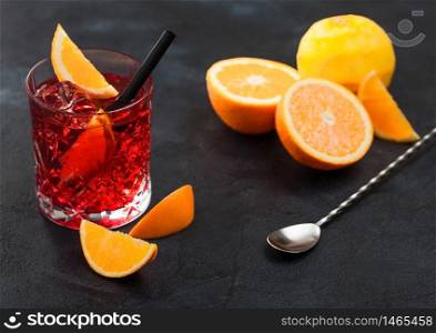 Negroni cocktail in crystal glass with orange slice and fresh raw oranges with spoon on black background. Top view