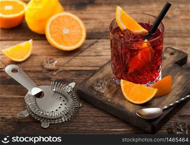 Negroni cocktail in crystal glass with orange slice and fresh raw oranges on chopping board with strainer on wood background. Top view
