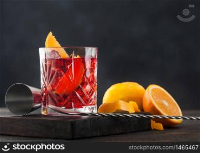 Negroni cocktail in crystal glass with orange slice and fresh raw oranges on chopping board with spoon on wooden background.