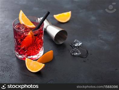 Negroni cocktail in crystal glass with orange slice and fresh raw oranges with jigger and ice cubes on black background. Top view