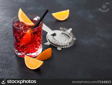 Negroni cocktail in crystal glass with orange slice and fresh raw oranges with strainer on black background. Top view