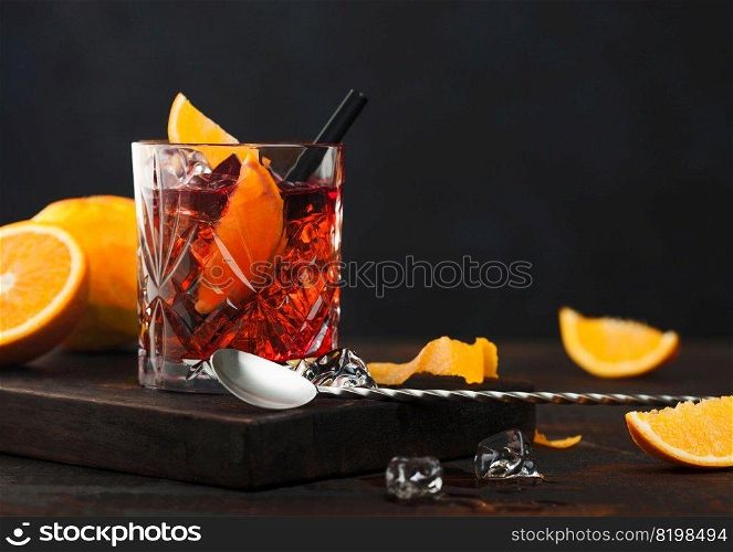 Negroni cocktail in crystal glass with orange slice and fresh raw oranges on chopping board with spoon on wooden background.