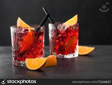 Negroni cocktail in crystal glasses with orange slice and black straw on black table background. Macro