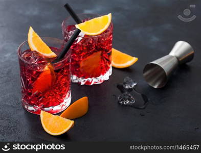 Negroni cocktail in crystal glasses with orange slice and fresh raw oranges with jigger and ice cubes on black background. Top view