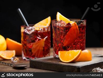 Negroni cocktail in crystal glasses with orange slice and fresh raw oranges on chopping board with strainer on wood background. Macro