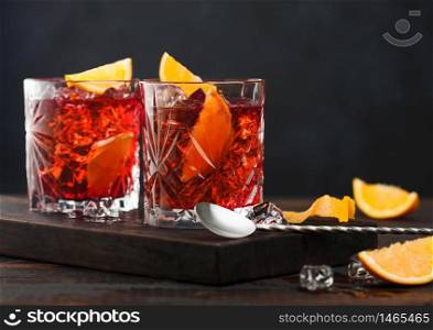 Negroni cocktail in crystal glasses with orange slice and fresh raw oranges on chopping board with spoon on wooden background.