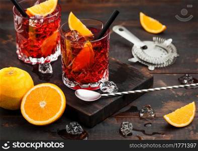 Negroni cocktail in crystal glasses with orange slice and fresh raw oranges with strainer and spoon on dark background.