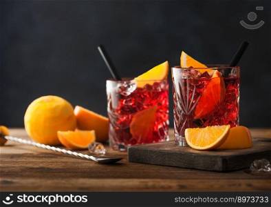 Negroni cocktail in crystal glasses with orange slice and fresh raw oranges on chopping board with strainer on wood background. Top view