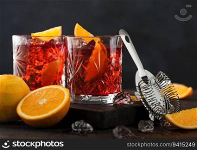 Negroni cocktail in crystal glasses with orange slice and fresh raw oranges on chopping board with strainer on wooden background.