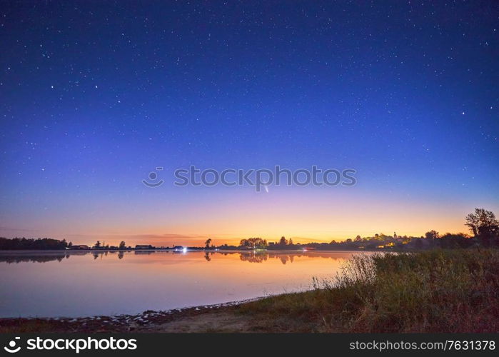 Neowise comet with light tail in night sky over lake Ozero with a beautiful orange sky at sunset or sunrise, Belarus.