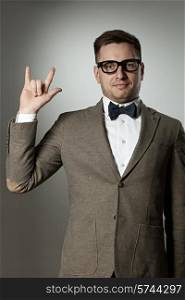 "Nerd in eyeglasses and bow tie showing "rock on" gesture against grey background"