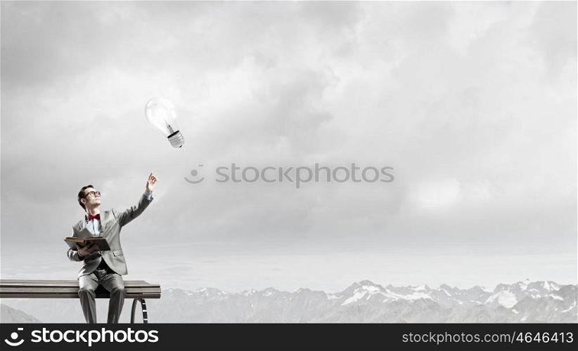 Nerd with book. Young businessman sitting on bench with book in hands and pointing with finger at light bulb
