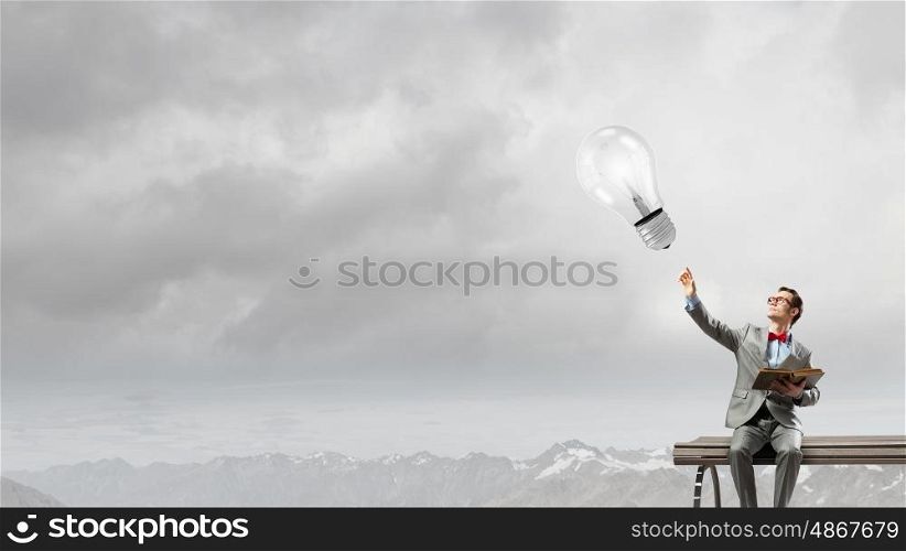Nerd with book. Young businessman sitting on bench with book in hands and pointing with finger at light bulb