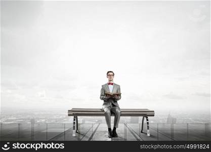 Nerd with book. Young businessman wearing red bow tie sitting on bench with book in hands