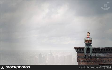 Nerd with book. Young businessman wearing red bow tie sitting on building top with book in hands