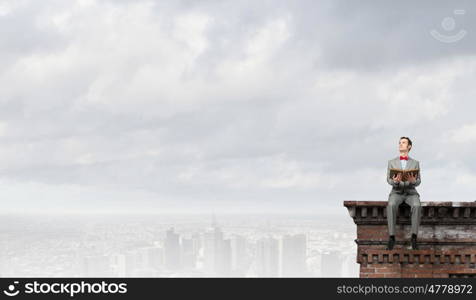 Nerd with book. Young businessman wearing red bow tie sitting on building top with book in hands