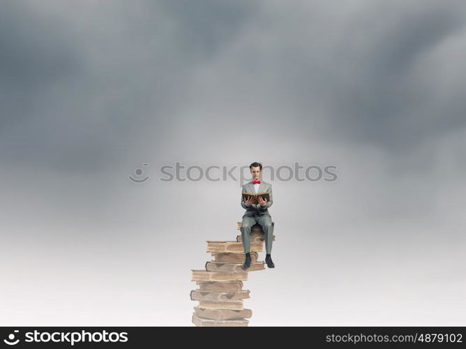 Nerd with book. Young businessman wearing red bow tie sitting on pile of old books