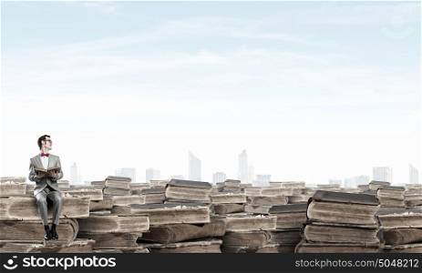 Nerd with book. Young businessman wearing red bow tie sitting on pile of old books