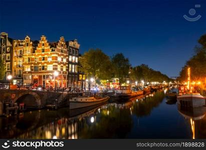 Netherlands. Calm on the night canal of Amsterdam. Residential barges and boats are moored. Reflection in the water of traditional houses and a bridge. Amsterdam Canal With Boats and Traditional Houses at Night
