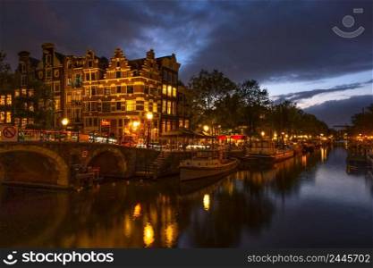 Netherlands. Cloudy evening on the canal of Amsterdam. Boats and houseboats are moored. Reflection in the water of traditional houses and a bridge. Embankment of the Amsterdam Canal on a Cloudy Evening