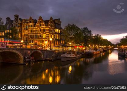 Netherlands. Cloudy evening on the canal of Amsterdam. Houseboats and boats are moored. Reflection in the water of traditional houses and a bridge. Amsterdam Canal at Cloudy Evening