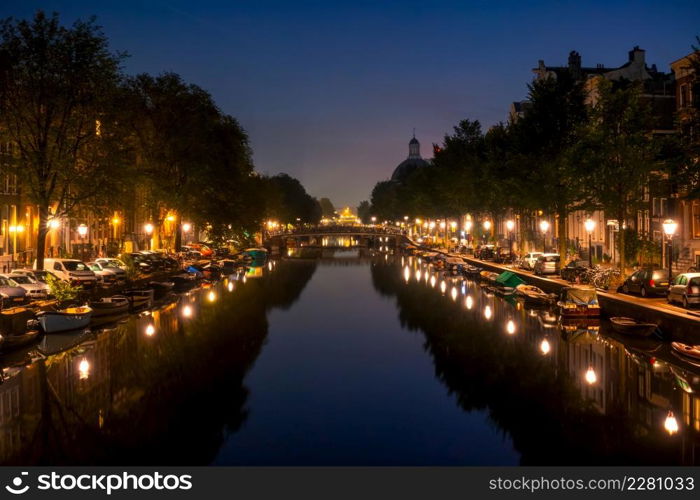 Netherlands. Night Amsterdam. Lanterns and parked cars on the embankments. Many boats are moored along the banks of the canal. Night Canal in Amsterdam With Parked Cars and Boats
