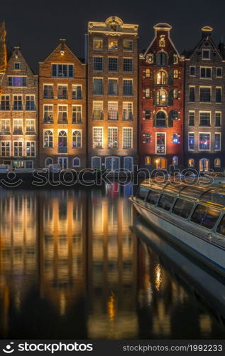 Netherlands. Night Amsterdam. Multi Colored windows of authentic houses near the water and a pleasure boat. Houses on Water in Night Amsterdam and Pleasure Boat