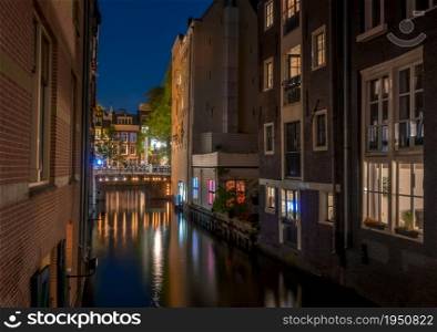 Netherlands. Night on the narrow canal between the Amsterdam houses and the illuminated bridge. Night Canal Between Amsterdam Houses