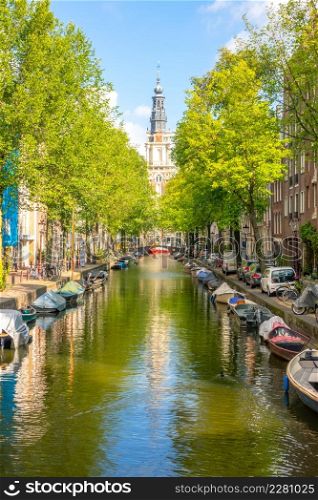 Netherlands. Sunny summer day on the Amsterdam canal. Parked cars on the embankment and many moored boats on the water. Cathedral building in the distance. Amsterdam Canal with Boats on a Sunny Day
