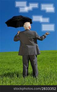 Network concept photograph of a businessman standing in a green field with an umbrella holding out his hand and looking at graphic screen boxes