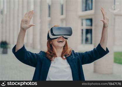 New age of business. Impressed ginger business woman testing virtual reality, using mobile VR headset with excited face expression, standing alone on city street in front of office buildings. Impressed ginger business woman testing virtual reality, using mobile VR headset outside