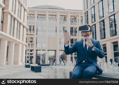 New age of business. Impressed young businessman or office worker testing virtual reality goggles, using VR headset with excited face expression, sitting alone outdoors on bench near fountains. Impressed young businessman or office worker testing virtual reality goggles, using VR headset