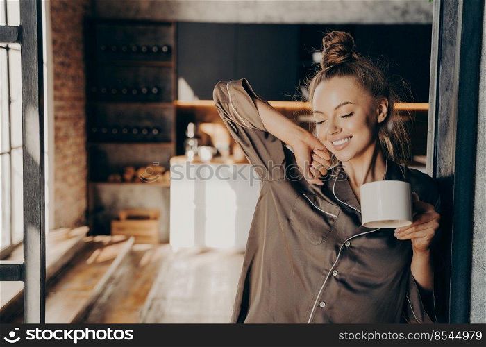 New day. Happy beautiful brunette female stretching with cup of coffee in her hand enjoying morning sunshine while standing in kitchen doorway in satin pajama. People and lifestyle concept. Happy beautiful brunette female stratching with cup of coffee in her hand while standing in kitchen