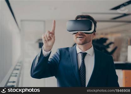 New experience. Smiling surprised bearded businessman in 3d goggles using augmented reality in work, dressed formally, pointing at object in user interface. Modern technology and business concept. Smiling bearded businessman in 3d goggles using augmented reality in work