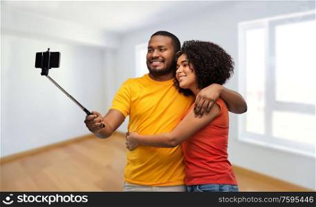 new home, real estate and people concept - happy african american couple taking picture by smartphone on selfie stick over empty apartment on background. african american couple taking selfie by cellphone